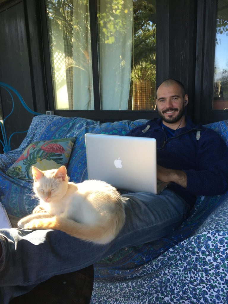 Jonathan Clarkin working on a laptop with Mally the cat sleeping on his legs while housesitting in Spain