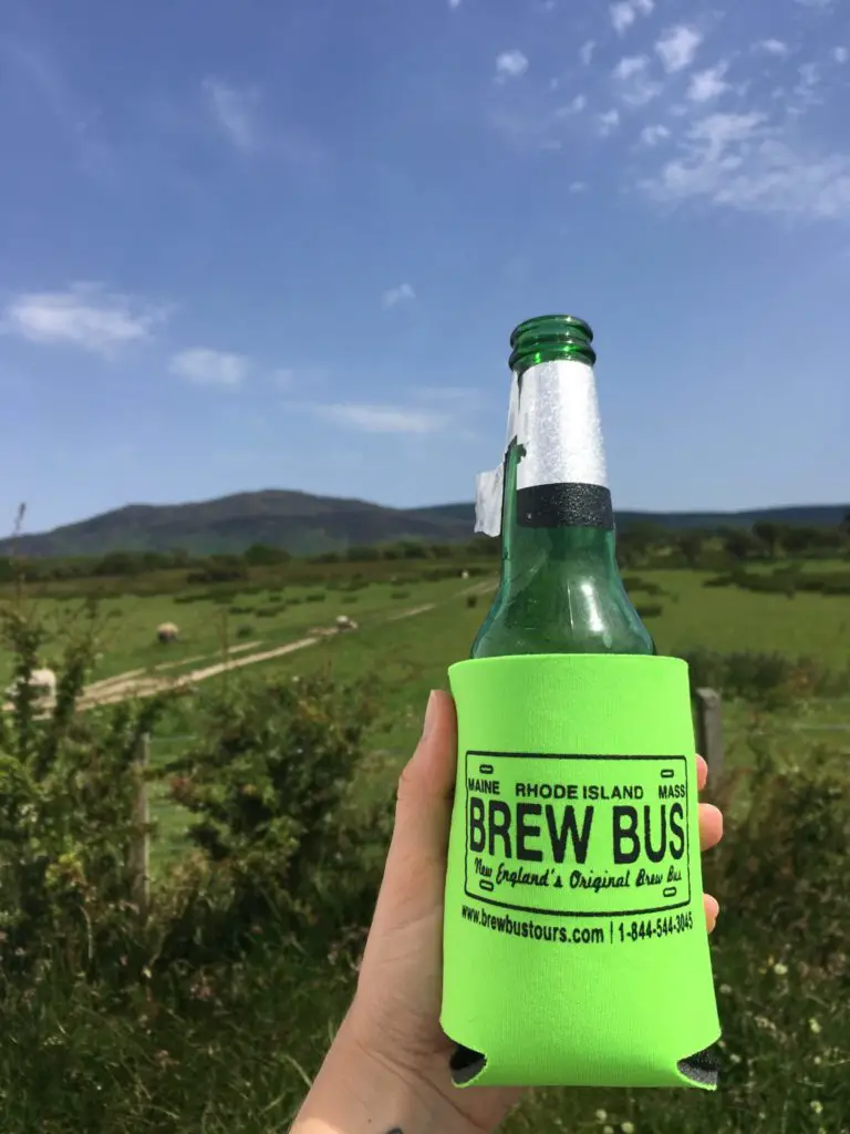 Maine Brew Bus coozie on a beer bottle held up against a blue sky and green fields on a visit to the Isle of Arran in Scotland
